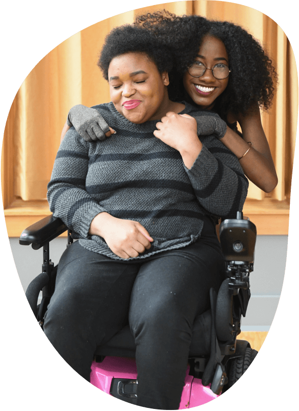 A Black woman in a black and pink power wheelchair has her eyes closed and is smiling while being hugged from behind by another Black woman who is wearing compression gloves. The two women have their hair tangled together from the embrace.