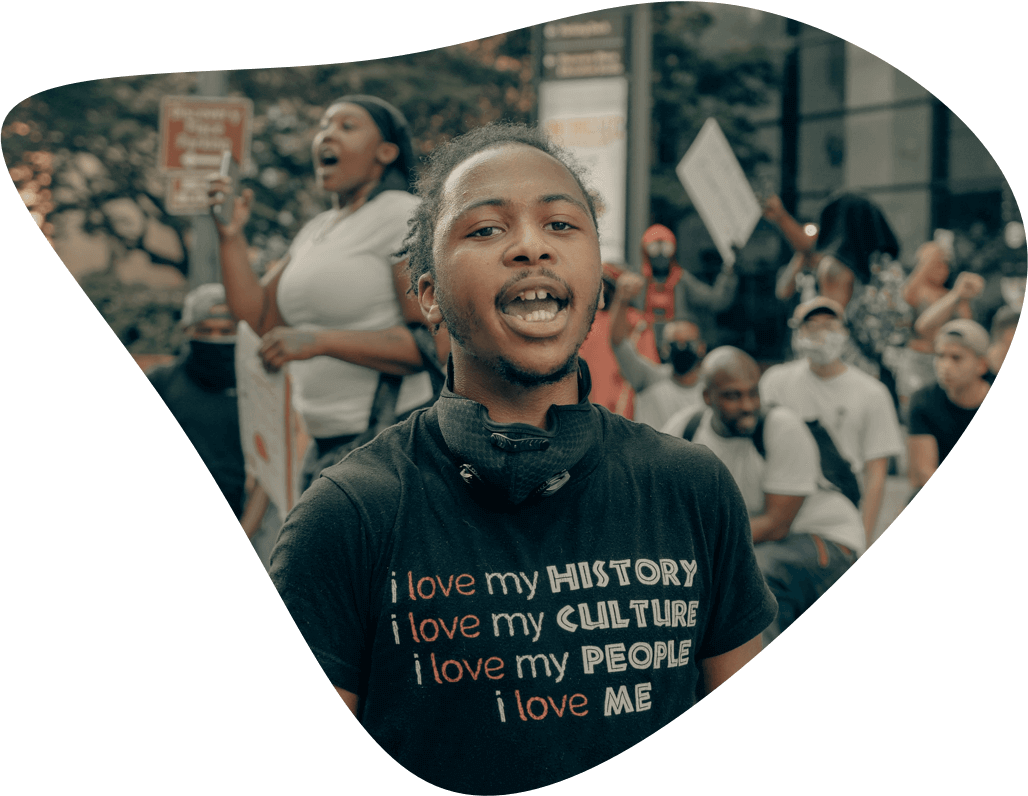 Young Black man stands on street during the daytime wearing a tee-shirt that reads “i love my history, i love my culture, i love my people, i love me”