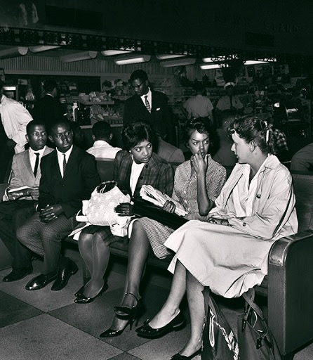 Three well-dressed Black women lean in to talk as they sit on a pew in a large space filled with people. Two young Black men sitting in the same pew look in their direction.