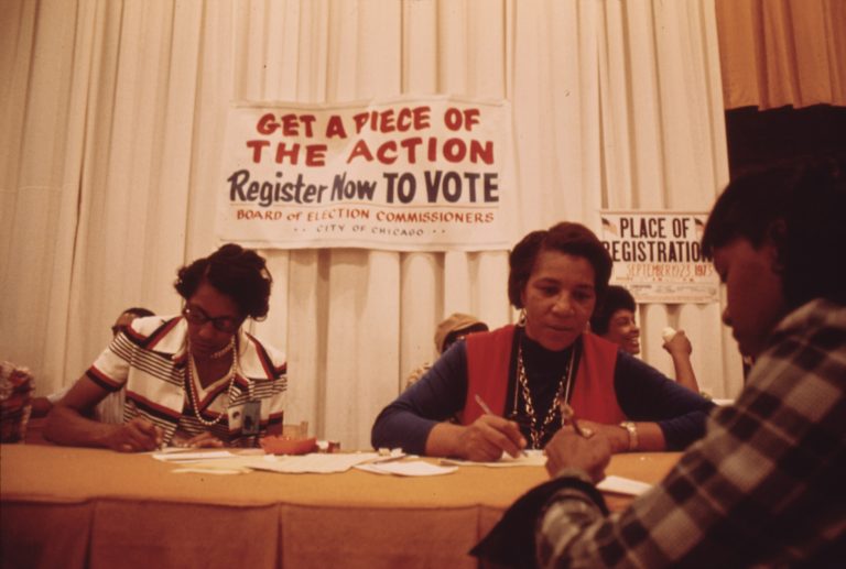 Three Black women sit at a table and write on pieces of paper. Above them signs read "Get a piece of the action. Register now TO VOTE. Board of elections commissioners. City of Chicago." and "Place of registration September 19-23 1973"