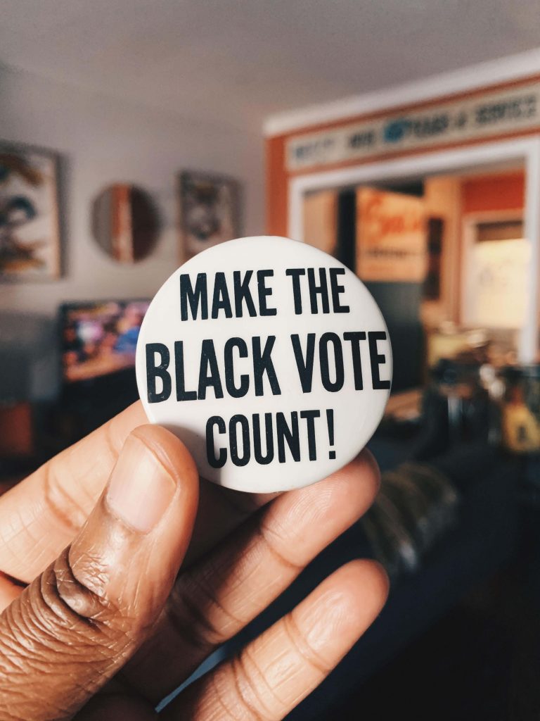 A hand holds up a 1970s white celluloid button that reads "Make The Black Vote Count" in black letters.
