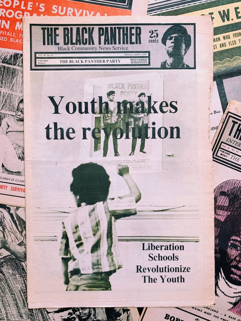 The front page of the Black Panther Party newspaper shows a Black child, back to camera, in a striped shirt raising a right hand to a poster of two men Black Panthers. Headlines on the front page includes "Youth makes the revolution. Liberation Schools Liberate the Youth."