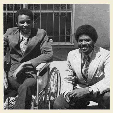 Disability activist and Black Panther Party member, Bradley Lomax, smiling and wearing a dark suit and patterned shirt, sits in a wheelchair to the left of his brother Glenn Lomax who squats beside Bradley in a light suit and tie.