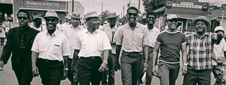 Martin Luther King Jr. and Stokely Carmichael walk side by side with a group of four other leaders and a couple folks walk with them, right behind. They walk down a road with the shop signs Southern Glass Co. and a grocer visible behind.