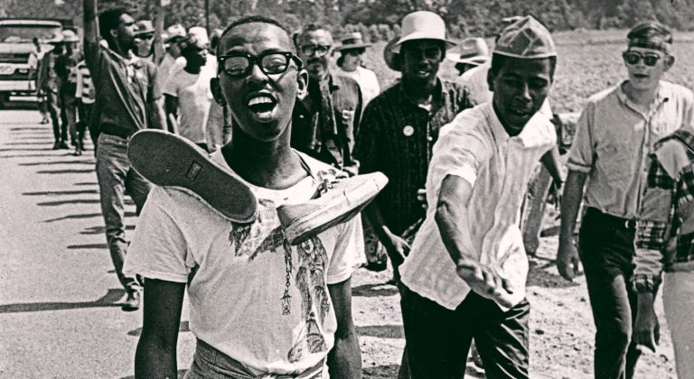 A young Black activist appears mid-sentence. He wears glasses, a printed teeshirt, light colored pants, and tennis shoes tied together over his shoulders. He walks with a line of almost twenty around and behind him along a rural road.