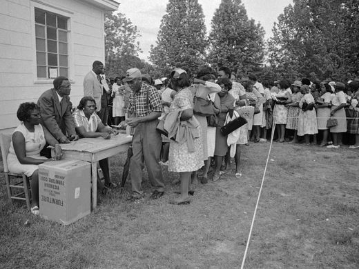 A long dense line of well-dressed Black voters stand before a table and behind a white string. At the front of the line, an elder submits a ballot to a few people who sit and stand behind the table.