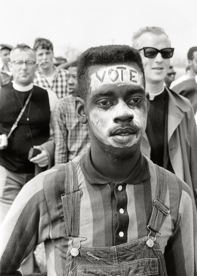 A Black man in overalls and a striped shirt marches with many protesters behind him. His face is painted white with the letters "VOTE" carved across his forehead.