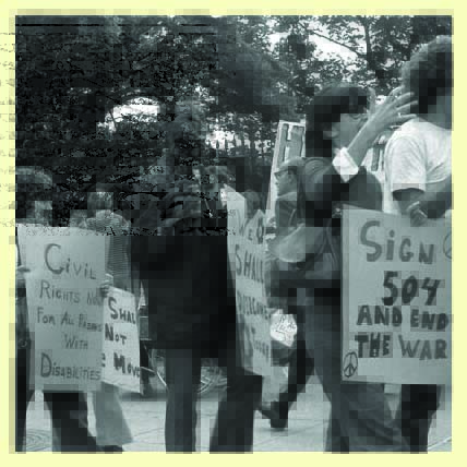 Five protestors march and chant holding signs that read "Civil rights now for all persons with disabilities" and "Sign 504 and end the war". At center a Black protester holds a sign that says "We shall overcome"
