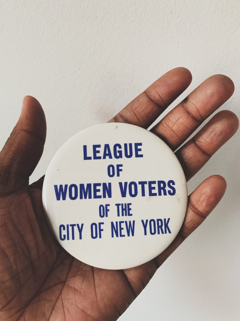 A hand holds a white button with blue lettering that reads "League of Women Voters of the City of New York"