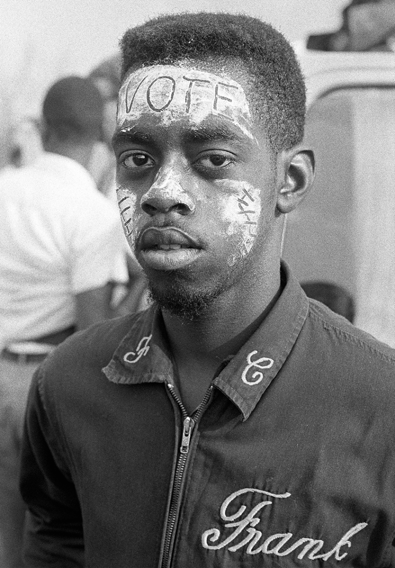 A Black man in a jacket with cursive lettering spelling "Frank" looks into the camera. His face is painted white with the letters "VOTE" carved across his forehead.