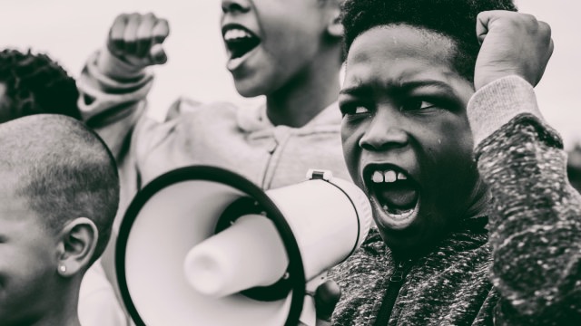A young Black boy yells into a megaphone and raises his left fist in the air. Around him are a few other smiling, exclaiming young children.