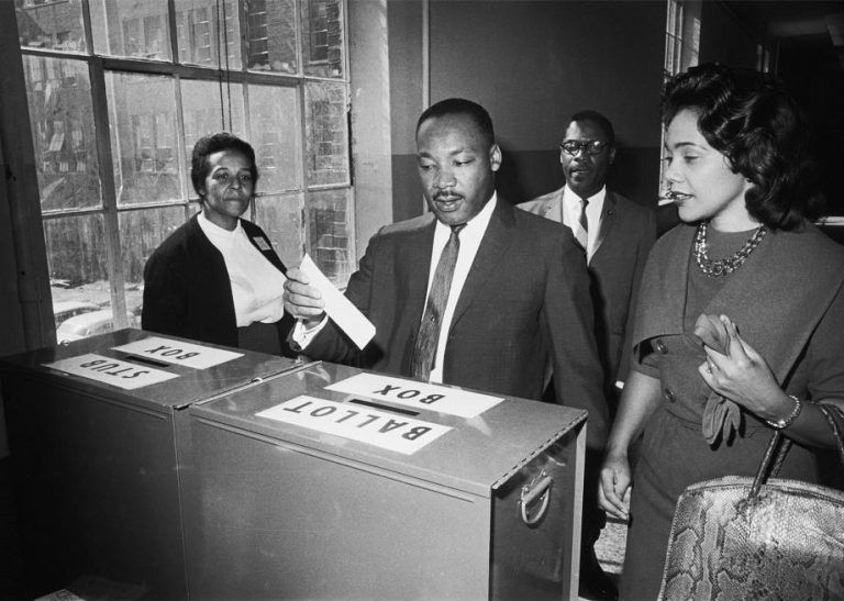 In front of two others, Martin Luther King Jr. and Coretta Scott King approach a labeled ballot box. Martin Luther King Jr., midsentence, motions to place his ballot in the box.