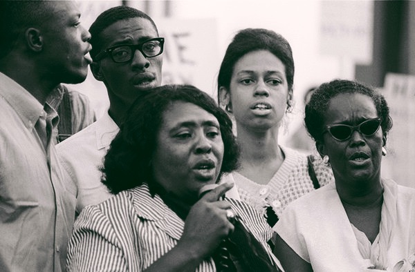 Five freedom riders stand together. One woman, in the front, speaks into a microphone.