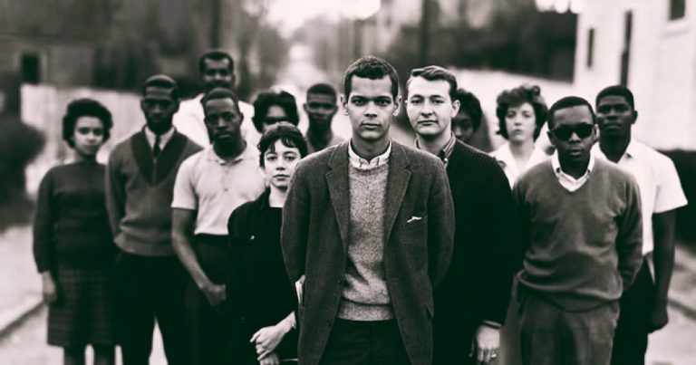 A young Black leader stands in focus the forefront of a well-dressed group of nine Black and three white young activists. They stand together in a cluster on a residential street, a few with their hands behind them.