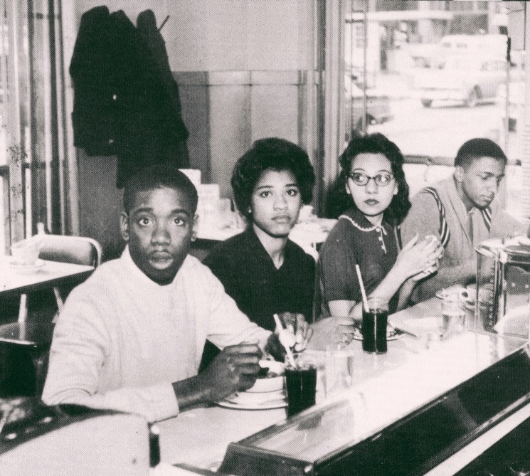 Four young Black activists sit at a lunch counter with sodas and meals in front of them. Two closest to the camera look into the camera. The other two look in other directions. The table behind them is empty and through the glass diner door and window cars whiz by.