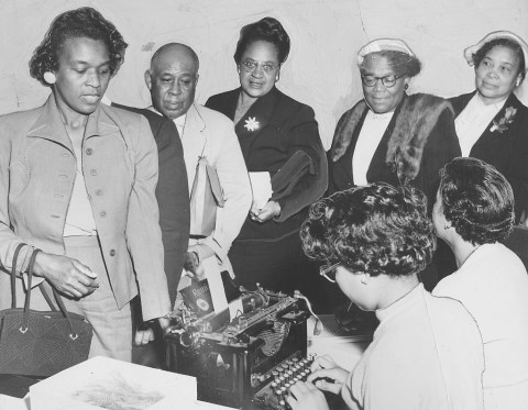 Five well-dressed Black elder voters line up in front of a two women typing on typewriters.