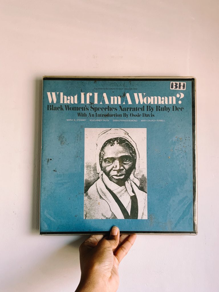 A hand holds a vintage record with an illustration of Sojourner Truth and text that reads "What If I Am A Woman? Black Women's Speeches Narrated by Ruby Dee With An Introduction by Ossie Davis. Maria W Stewart. Sojourner Truth. Sarah Parker Remond. Mary Church Terrell."