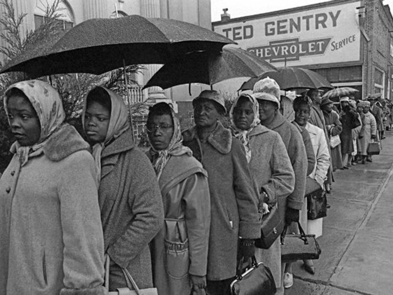 Holding umbrellas, a long line of Black women voters in overcoats and kerchiefs stand in the rain on a sidewalk.
