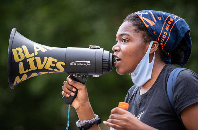 Young Black woman wearing blue and orange headkerchief, white face mask around her chin and black Tee holding black bullhorn reading Black Lives Matter in yellow words.