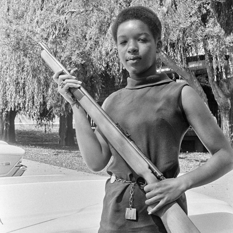 A young Black woman in a dress holds a rifle and looks into the camera lens.