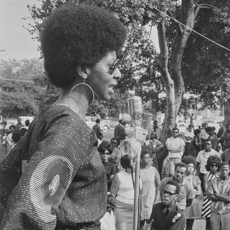 A woman Black Panther member addresses an on-looking crowd through a microphone. She wears sunglasses, large hoop earrings, and a patterened blouse.
