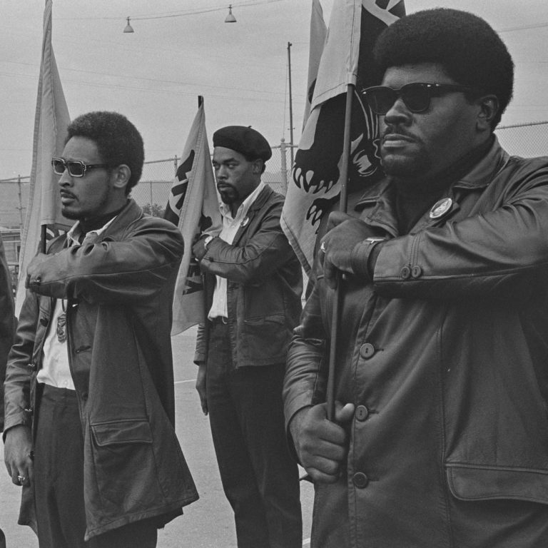 Three men Black Panther members in leather jackets, some wearing sunglasses, hold flags to their right.