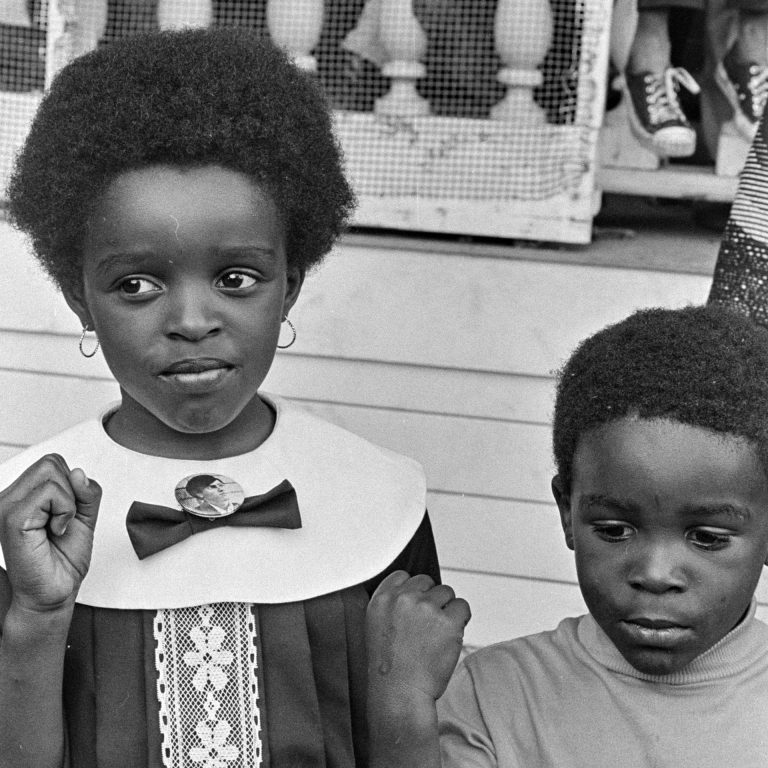 Two young Black children raise their right fists in the air. One wears a Huey Newton button pinned to the bow of her dress' lapel.
