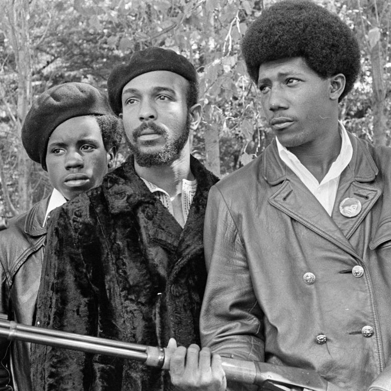 Three men Black Panther members stand by. They wear white shirts under leather or fur jackets, and two wear black berets. The Panther in the foreground holds a rifle.