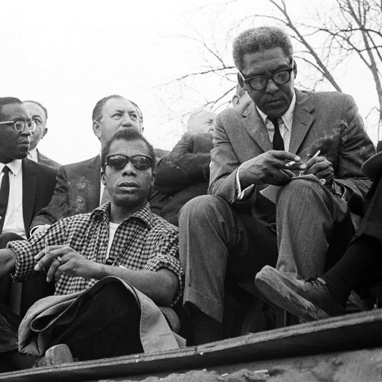 James Baldwin and Bayard Rustin sit to the front of a group of men. Baldwin, in a checkered shirt, smokes. Rustin, in a suit, writes in a notebook with his left hand and holds a cigarette in his right hand.