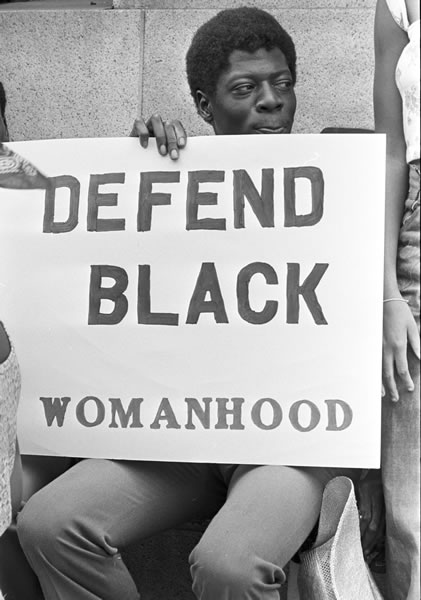A Black man sits and holds a sign on his lap that says "Defend Black Womanhood". In supporting Joan Little, he sits outside the couthouse where Little's trial was held, in North Carolina, in 1975.