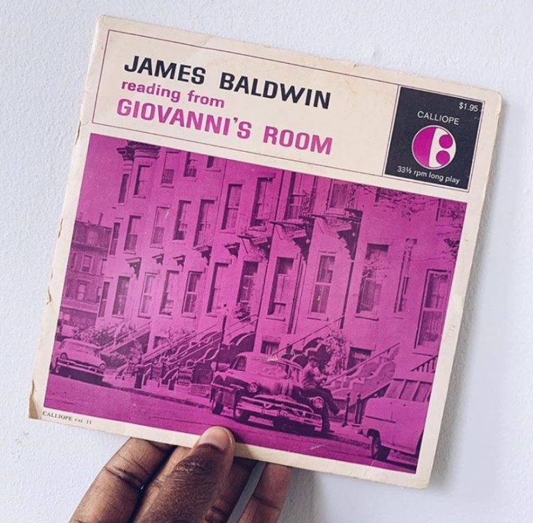 A hand holds a vintage record with a picture of a street with brownstone homes and parked cars and text that reads "JAMES BALDWIN READING FROM GIOVANNI'S ROOM"