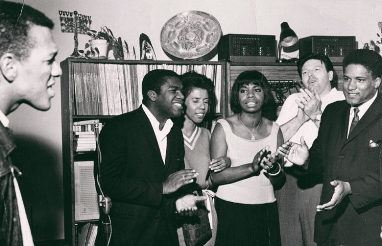 Nina Simone stands alongside four people and they clap and sing along in admiration of a man to their right who sings passionately. Behind the group is a shelf stacked with records, books, a stereo system, a menorah, and other decor.