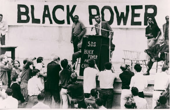 A Black activist leader stands at a podium in front of an enormous "BLACK POWER" banner on stage with a few others and in front of a crowd of listeners.