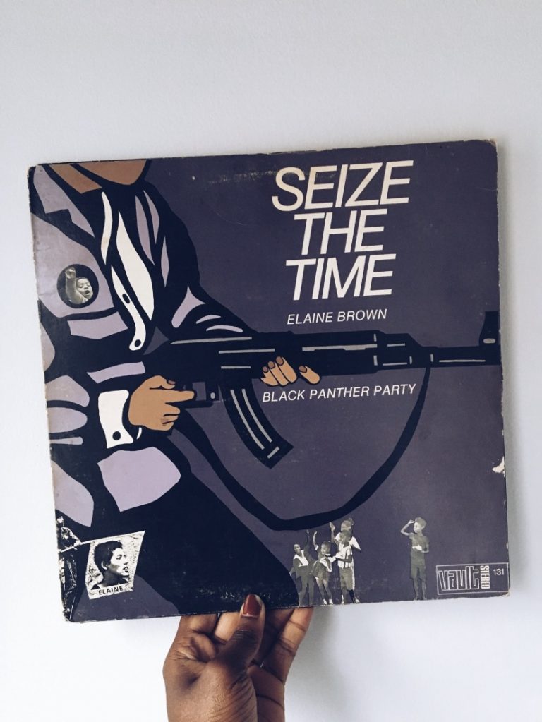 A hand holds a vintage record with a color illustration of a figure wearing a white shirt, grey jacket, and black pants and holding a rifle. On the figure's lapel is a button showing a toddler raising a fist in the air. Around the figure are small photographic collages of Black children waving and saluting. Text on the record reads "SEIZE THE TIME. ELAINE BROWN. BLACK PANTHER PARTY."