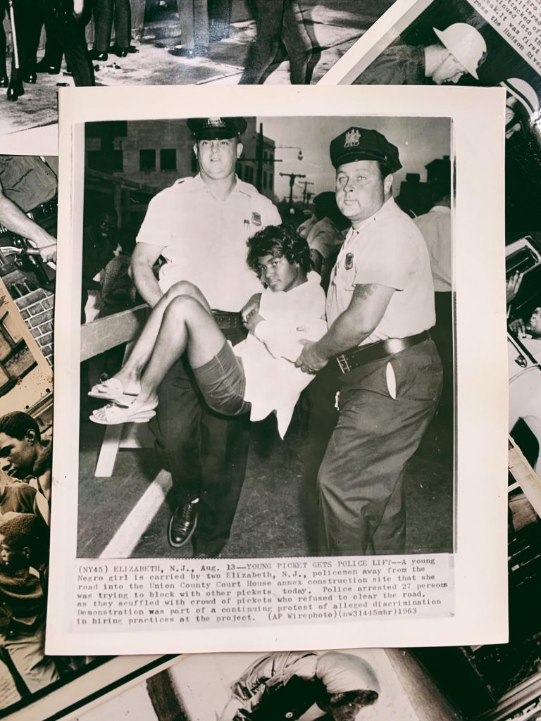 A vintage photo shows two white police officers carrying a young Black woman protester by her arms and legs. Under the photo is a caption that explains the woman protestor was blocking a road in Elzabeth, NJ on August 13 and was arrested with 27 other picketers.