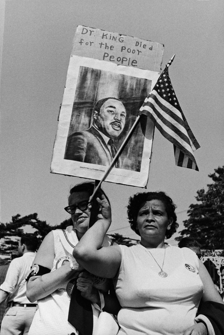 Two women hold up an American flag and a sign with a painting of Martin Luther King Jr. on it that says "Dr. King dies for the poor people".