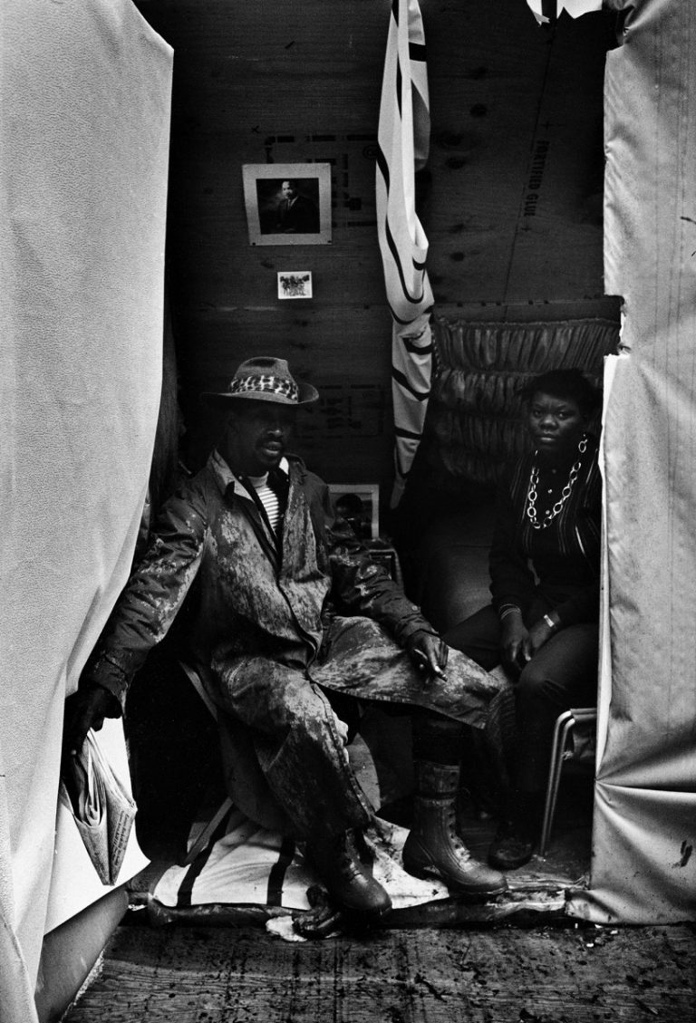 A Black man, wearing a hat and overcoat and holding a newspaper, and woman wearing a large necklace, sit together. Above them in their encampment is a photograph of Martin Luther King Jr. and tall fabric dividers.