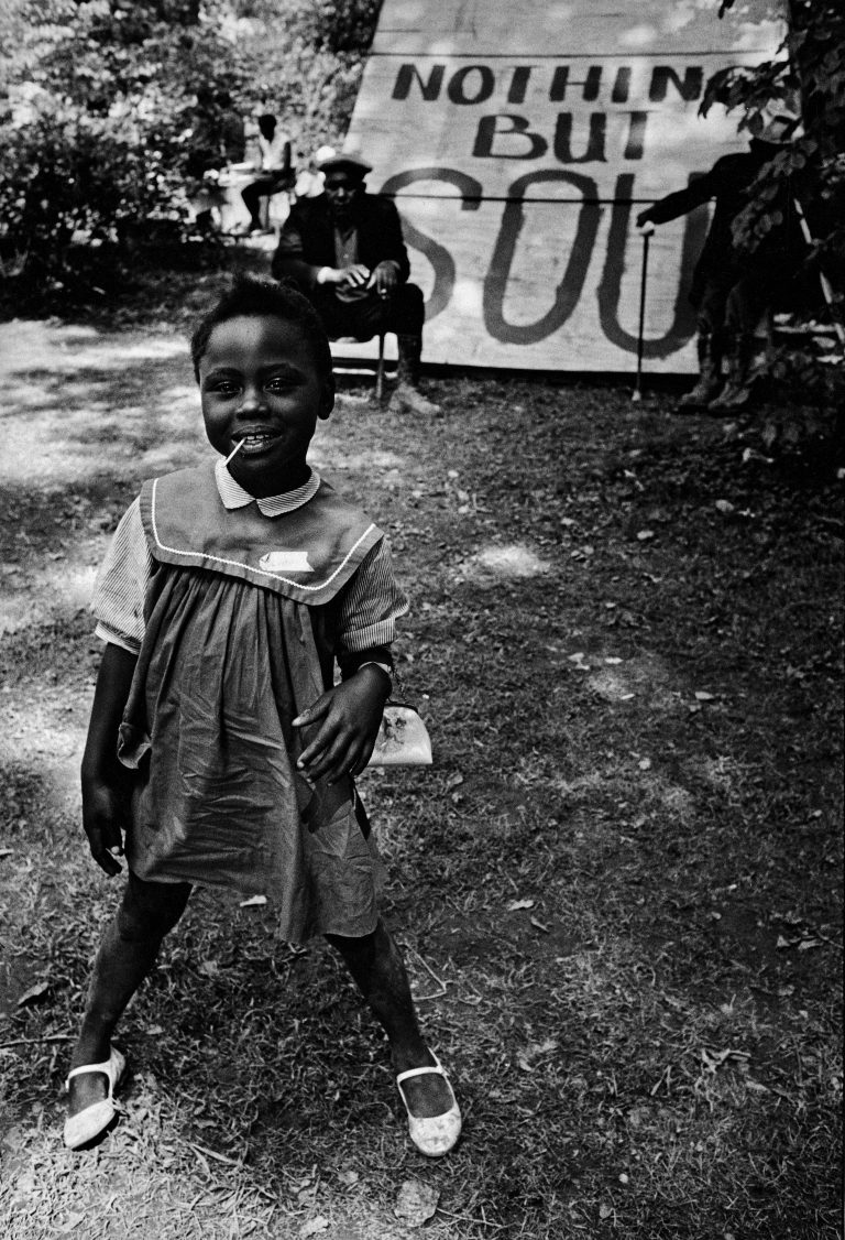 A young Black girl wearing a dress, dress shoes, and a small purse smiles at the camera with a lollipop in her mouth. Behind her two Black elders sit by a large sign that says "Nothing but SOUL"