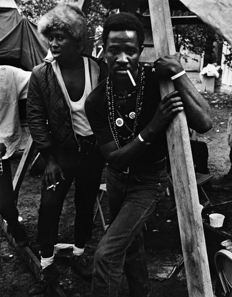 Two Black men protestors stand by a structure of an encampment. Smoking cigarettes, they look into camera lens.