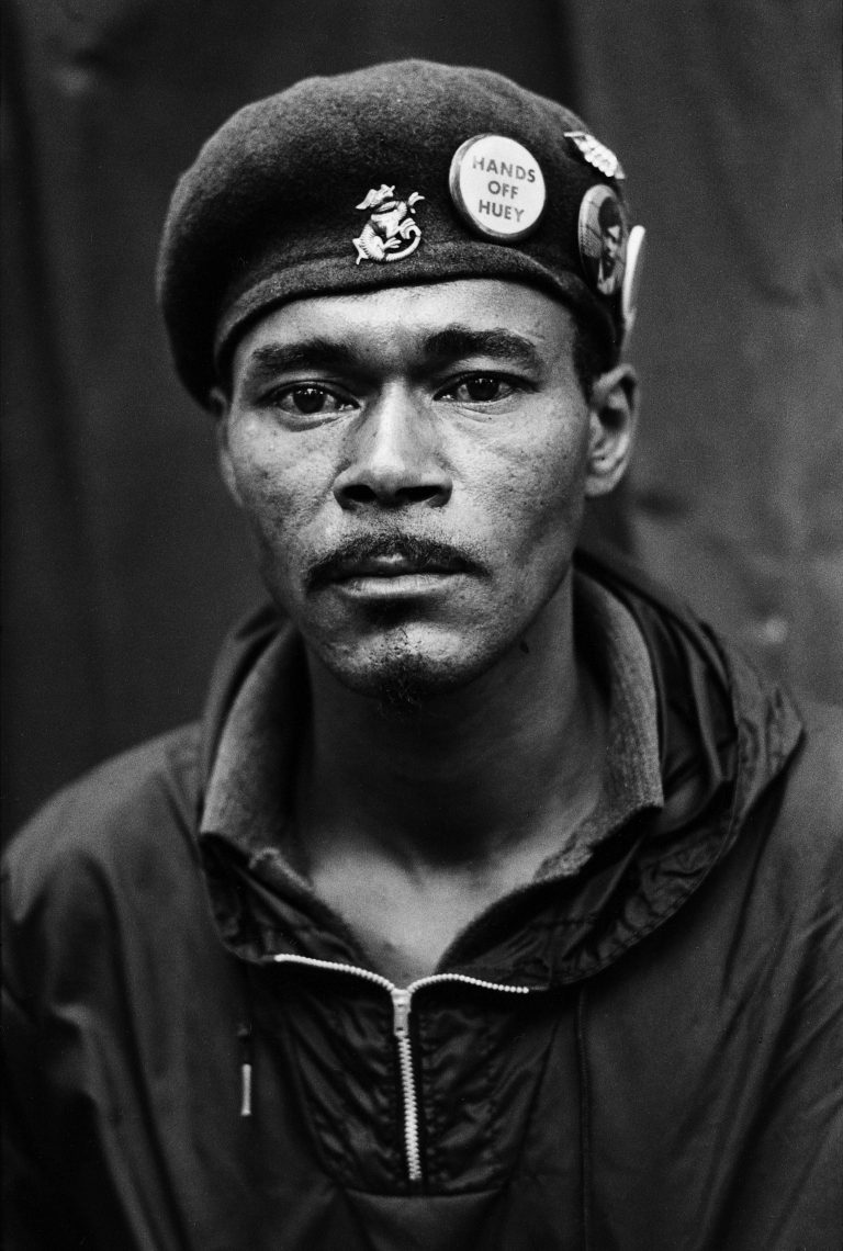 A Black man protestor looks into the camera lens. He wears a rain pullover and a beret with several buttons including one that says "HANDS OFF HUEY".