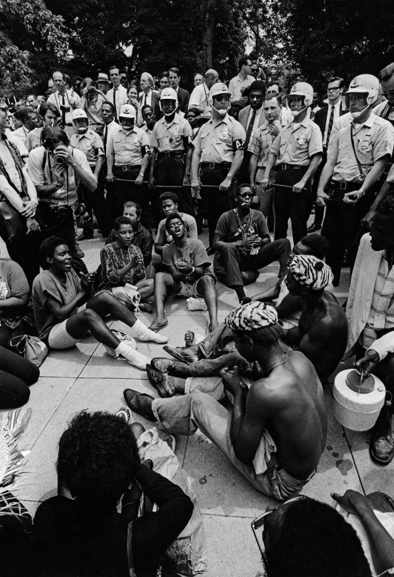 A group of Black protestors sit in a small circle on the ground, singing and clapping. They are surrounded by white police officers in uniform and riot gear, onlookers, and photographers.