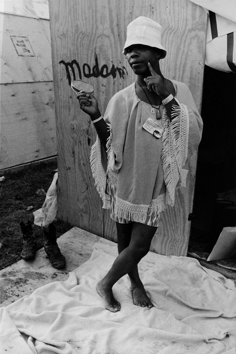 A Black queen in a white hat and fringed dress poses with a mirror in one hand and the other hand pointing to their cheek. In front of their encampment, they stand barefoot next to a pair of cowboy boots. Behind them, painted words on wood spell "Madam".