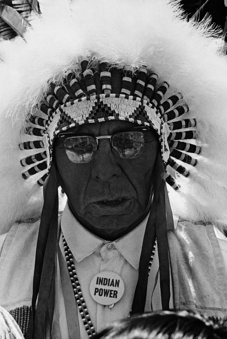 An indigenous elder looks into the camera lens mid-sentence. He wears glasses, a beaded, ribboned, and feathered headdress, and a button on his collared shirt that reads "INDIAN POWER".