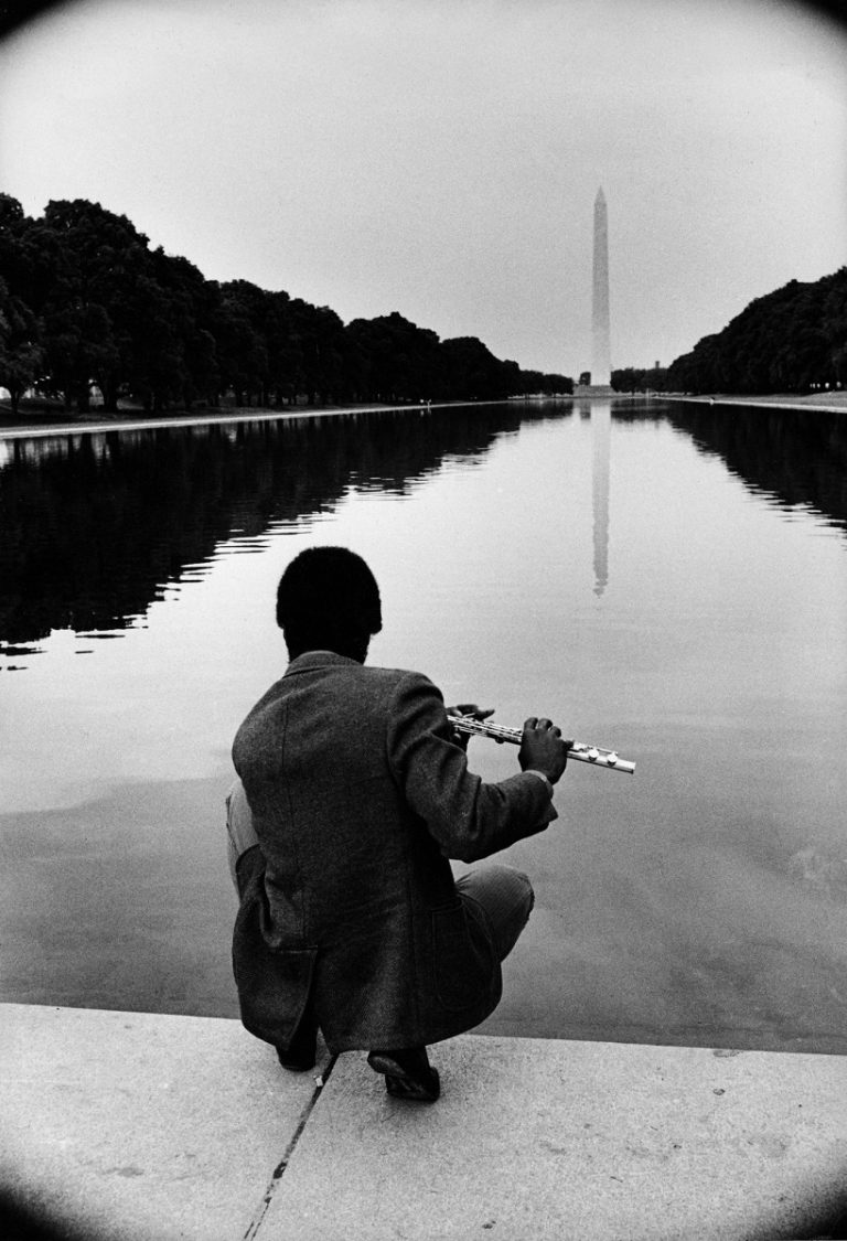 A Black man plays a flute, his back to the camera, sitting at the edge of a pool water that stretches to the Washington Monument in the distance.