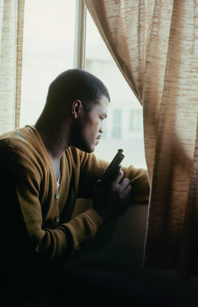A man in a taupe cardigan pulls back a beige semi-opaque curtain to look out the window. He holds a gun in his right hand.