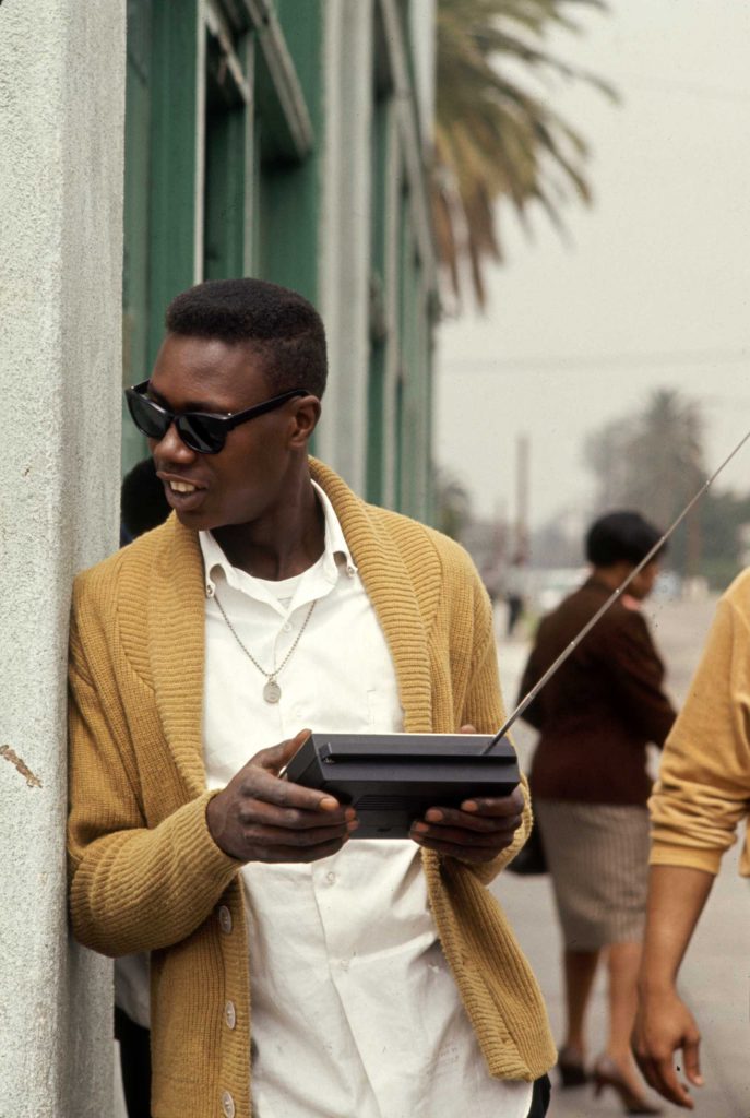A young Black man in a yellow cardigan, white shirt, metal necklace and sunglasses leans against a building facade and holding a radio, looks to his right.