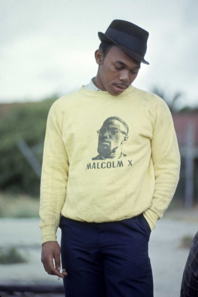 A Black man wears a yellow pullover with Malcolm X's portrait and "Malcolm X" written on it. He wears a dapper hat and smokes a cigarette.