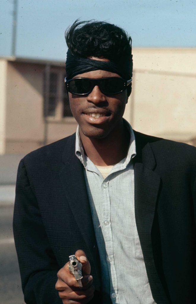 A smiling man in sunglasses, head wrap, and blazer holds a gun.
