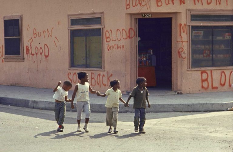 Four small children cross a street holding hands and looking to their left. Behind them, a peach colored corner store facade is covered in spray painted words including burn, blood, blood brother, M+R.