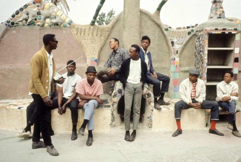 A group of nine young Black men dressed sharply sit on the steps of a concrete sculptured building with colorful mosaic decor. One man in a yellow cardigan and sunglasses holds a radio with a long antennae.